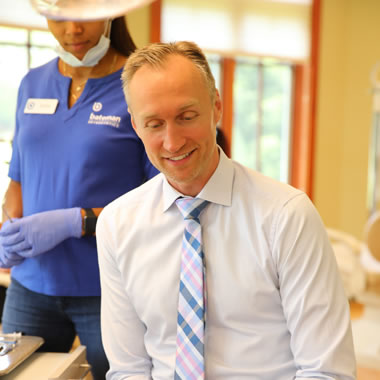 Professional man smiling in orthodontic office with dental staff member wearing gloves in background.