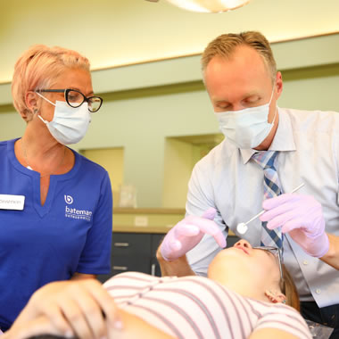 Male orthodontist and female assistant examine patient's teeth in clinical treatment room.