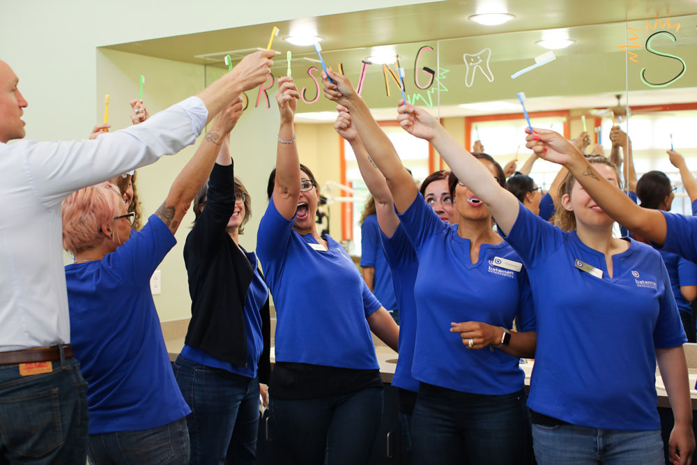 Team members in blue shirts celebrate together with raised hands in orthodontic office.