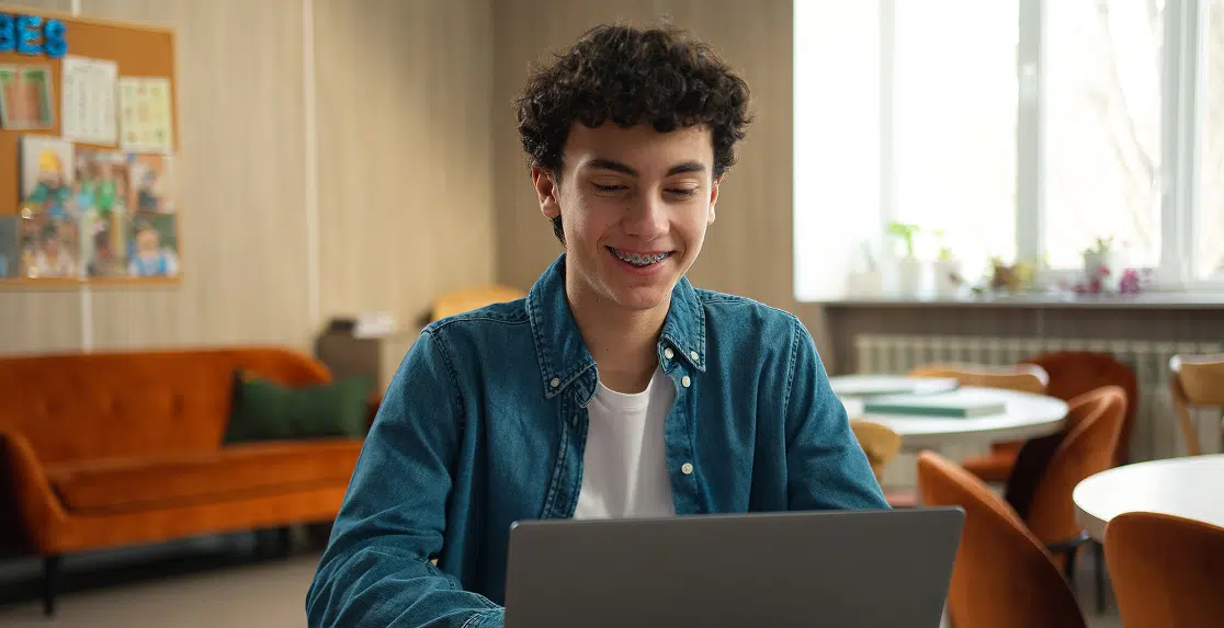 Smiling teenage boy with braces using laptop in bright, modern classroom setting.