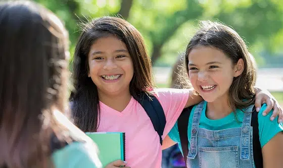 Three smiling teenage girls standing together outdoors in casual clothing, enjoying a friendly moment.