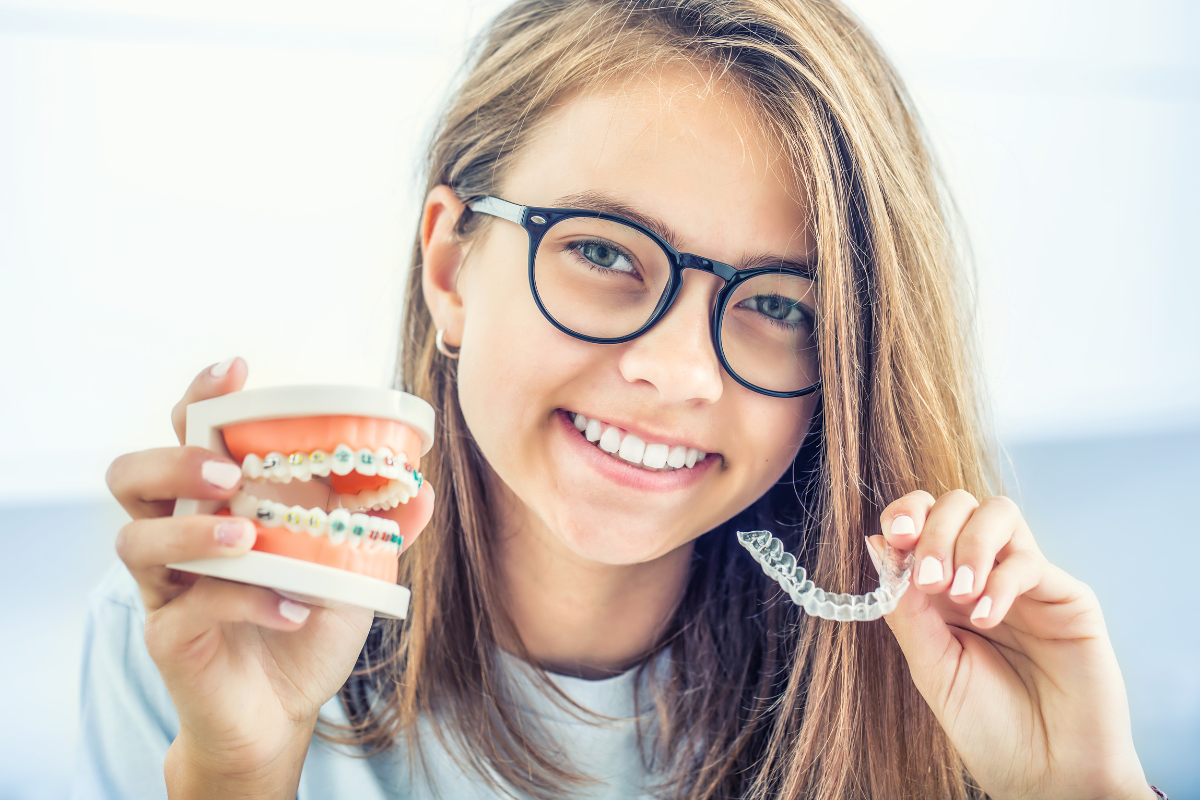 Smiling girl with glasses holding dental braces model and clear aligner tray.